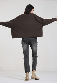 Brown oversized sweater with dropped shoulders, ribbed hem, paired with faded black jeans and beige ankle boots, featuring buckled accents.