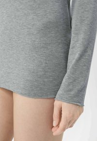 Chemise grise à manches longues en tissu doux et texturé avec un ourlet arrondi. Coutures visibles accentuant les poignets et l'ourlet.