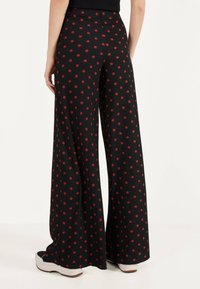 Kankaita leveitä housuja, jotka on valmistettu teksturoidusta mustasta kankaasta, jossa on punaisia pilkkuja. Vyötärö on korkea ja helma ulottuu kenkien päälle.