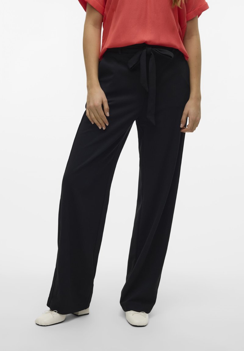 Pantaloni neri a gamba larga con cintura a cravatta in vita, caratterizzati da una texture liscia e un design fluido. Abbinati a una maglietta rossa a maniche corte.