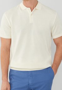 Ljus krämfärgad polo shirt i strukturerad tyg, med en tvåknappluck och korta ärmar. Bärs med blåa byxor.