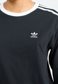 Černé tričko Adidas s dlouhým rukávem, bílým límečkem a pruhy na ramenou, s logem Adidas trefoil na levé straně hrudi.