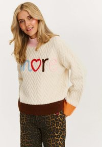 Dzianinowy kremowy sweter o wzorze w jodełkę z wielokolorowym napisem "więcej". Posiada różowy kołnierz i kontrastujące rękawy w odcieniach brązu i pomarańczy.