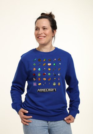 Niebieska bluza z pixelowanymi przedmiotami z Minecrafta w różnych kolorach, z napisem "MINECRAFT" wydrukowanym wyraźnie na dole. Miękka tkanina, luźny krój.