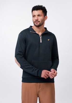 Pulóver verde oscuro, texturizado, con cuello con cremallera. Presenta un panel contrastante en color beige en la manga y un pequeño logo dorado en el pecho.