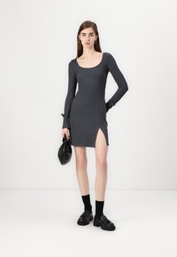 Grå långärmad klänning med figursydd silhuett, sido-slits och strukturerad tyg. Modellen håller en svart väska och bär svarta strumpor och skor.