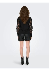 Top negro con mangas transparentes y texturizadas; combinado con pantalones cortos de cuero sintético negro y botines negros. Vista trasera, cabello rizado visible.