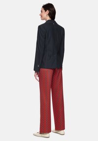 Navy tailored jacket with gold stitching details, paired with wide-leg red patterned pants featuring geometric shapes. Worn with light-colored flats.