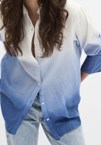 Kvinna med långt vågigt hår som bär en blå-vit gradient knappad skjorta över jeans, med en skymt av en brun topp under.