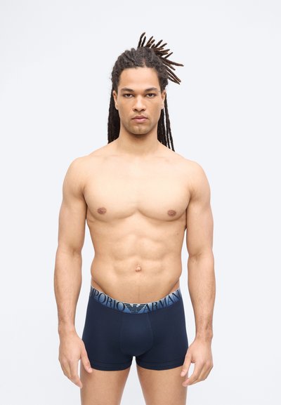 Άνδρας χωρίς πουκάμισο με μακριά dreadlocks στέκεται γυρισμένος προς τα εμπρός, φορώντας ναυτικό μπλε μποξεράκια Emporio Armani σε απλό ανοιχτό φόντο.