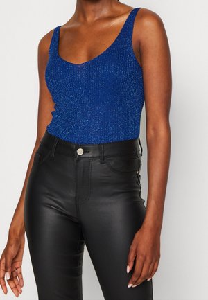 Vrouw met een sprankelend blauw mouwloos topje en een zwarte hoge taille leren broek, staand tegen een eenvoudige lichte achtergrond.