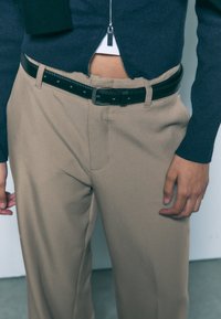 Beiget räätälöidyt housut, joissa on sileä pinta, musta nahkavyö ja laivastonsininen vetoketjullinen neulepusero, joka paljastaa osittain valkoisen topin alla.