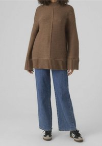 Brunstickad tröja med vida ärmar och ribbad nederkant, kombinerad med blå vida jeans och svarta sneakers med vita detaljer.