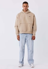 Sudadera beige de gran tamaño con bolsillo canguro y logo negro, combinada con vaqueros holgados azul claro con detalles de costuras curvas y zapatillas blancas.