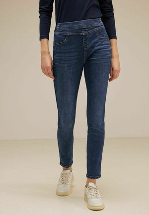 Persoon die donkerblauwe slim-fit jeans, een marineblauw shirt met lange mouwen en witte sneakers draagt, staand tegen een neutrale achtergrond.