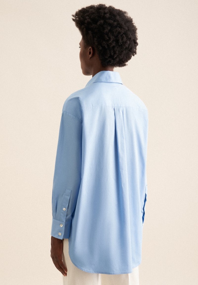 Personne aux cheveux courts et bouclés portant une chemise ample bleu clair boutonnée, vue de dos sur un fond beige.