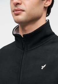 Μαύρο φλις μπουφάν με ψηλό γιακά, φερμουάρ μπροστά και ένα λευκό λογότυπο πουλιού στη αριστερή πλευρά. Μαλακή υφή και κανονικός σχε διασμός.