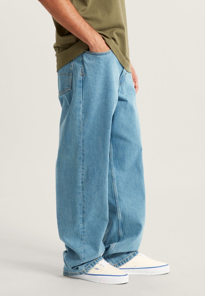Personne portant un jean bleu clair ample, une chemise vert olive, avec la main droite dans la poche et des baskets blanches à semelles bleues.