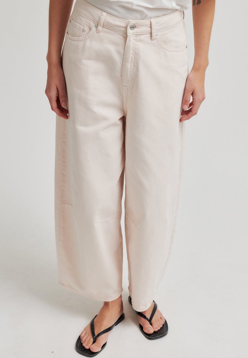 Personne portant un pantalon ample taille haute de couleur beige clair, associé à des sandales noires à tongs sur un fond clair.