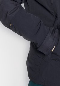 Svart, isolerad jacka med en strukturerad yta, elastiska ärmslut och en synlig varumärkesskylt på ärmen. Frampocket med dragkedja.