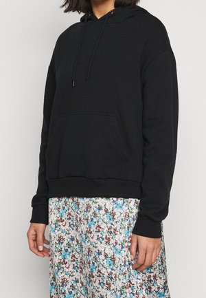 Person iført en enkel sort hættetrøje med snore og et blomsterprintet skørt med blå, røde og grønne blomster på en hvid baggrund.