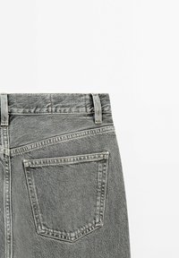Partie arrière d'un jean en denim gris montrant la ceinture, les passants de ceinture et une unique poche arrière sur fond blanc.