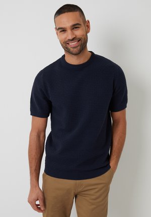 Navy knitted short-sleeve sweater with a textured pattern, round neckline, and ribbed cuffs, paired with light brown trousers.