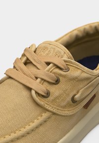 Κοντινή λήψη σε ανοιχτό καφέ καμβαδένιο casual παπούτσι με ανοιχτό καφέ κορδόνια δεμένα σε μεταλλικά ρέματα και κεντημένες λεπτομέρειες στην περιοχή των δακτύλων.