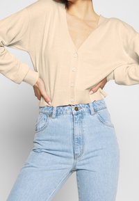 Beige v-ringad kofta med långa ärmar, med fem knappar fram och ribbad fåll, kombinerad med ljusblå högmidjade jeans.