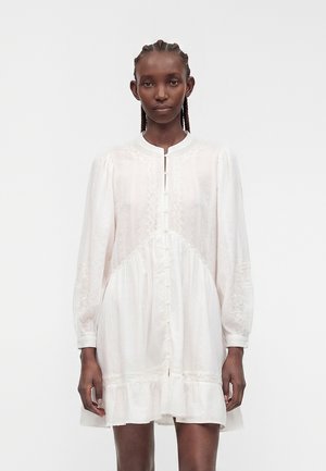 Femme à la peau foncée avec des cheveux tressés porte une mini-robe blanche texturée à manches longues avec une patte de boutonnage devant et un ourlet à volants, debout sur fond blanc.