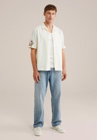 Camisa de manga corta de color claro con bordados florales, combinada con unos vaqueros de corte relajado de color azul claro y zapatillas blancas.