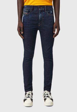 Mężczyzna ubrany w ciemnoniebieskie dopasowane dżinsy, żółty top i czarno-białe sneakersy, stojący na jednolitym jasnym tle.