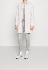 Gabardina de color beige claro con cierre de botones, dos bolsillos frontales, llevada sobre una camiseta gris. Unos vaqueros grises y zapatillas blancas completan el conjunto.