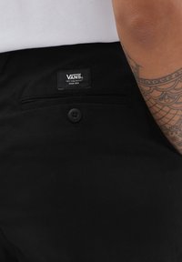 Sorte bukser med en Vans-label på taljen, en knaplukning og en sidelomme. Stoffet har en glat tekstur.