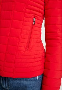 Gros plan d'une veste matelassée rouge avec une poche zippée verticale et un jean en denim bleu visible en dessous.
