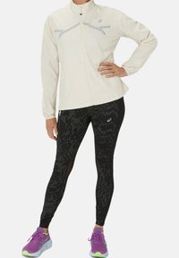 Persoon in een crèmekleurige zip-up jas, zwarte legging met patroon en gaaspanelen, en paarse hardloopschoenen, staande met één hand op de heup.
