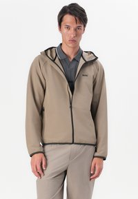 Beige, lichtgewicht jack met capuchon, zwarte accenten en een volledige rits. Draagt over een grijze, gestructureerde polo en gecombineerd met beige broek.