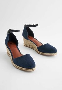 Scarpe a zeppa in suede blu navy con punta chiusa, cinturino alla caviglia e piattaforma in juta intrecciata. Superficie testurizzata e dettagli beige vicino alla suola.