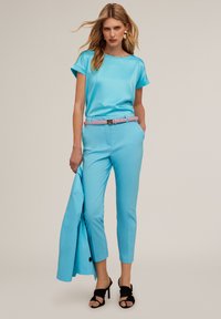 Ljust blå satin topp med uppvikta ärmar, matchande ljusblå skräddarsydda byxor, rosa bälte och svarta strappy klackar; modellen håller en blå kavaj.