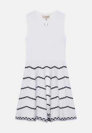 Robe sans manches blanche avec un haut ajusté. La jupe présente des motifs en zigzag noirs et des accents décoratifs. Tissu en coton lisse.