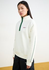 Kremkrāsas vilnas džemperis ar augstu apkakli, zaļu rāvējslēdzēju un logo. Iezīmes kontrastējošas zaļas sānu svītras un brīva piegriezuma siluets.