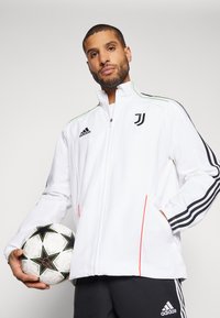 Chaqueta blanca con cremallera y franjas y acentos negros, que presenta el logotipo de la Juventus. El material parece suave, combinada con pantalones negros y un balón de fútbol.