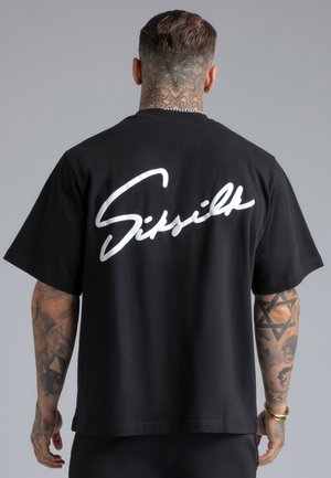 Mężczyzna z tatuażami na szyi i ramionach, ubrany w czarną koszulkę z dużym białym napisem „Siksilk” na plecach, na szarym tle.