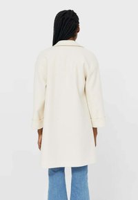 Cream-colored long coat with rolled three-quarter sleeves, structured shoulders, and a straight silhouette, paired with light blue flared jeans.
