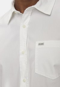 Camicia bianca con bottoni realizzata in tessuto liscio, presenta una tasca sul petto sinistro con etichetta nera "GUESS" e un colletto classico.