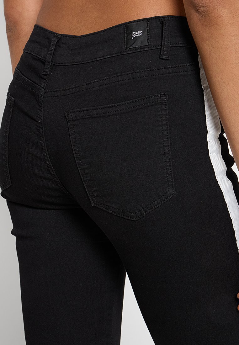 Jeans skinny noirs avec une bande blanche sur le côté, une poche arrière et une étiquette de ceinture de marque. Le tissu semble lisse avec un léger stretch.