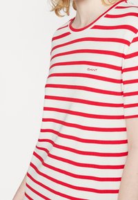 Personne portant un t-shirt blanc avec des rayures rouges horizontales et un petit logo "GANT" rouge sur la poitrine, coupé au niveau du cou et des bras.