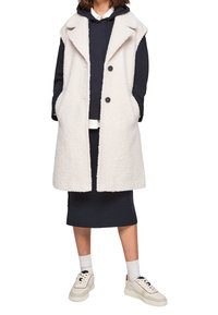 Un gilet di un colore crema, con una texture, colletto a scialle, bottoni frontali e tasche laterali, indossato sopra una felpa blu navy e una gonna. Sneakers bianche incluse.