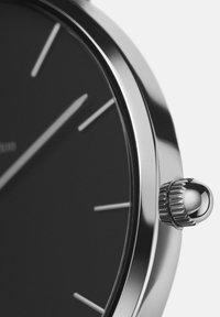 Apvalus nerūdijančio plieno laikrodis su juodu ciferblatu, sidabriniais valandų žymekliais ir tekstūruota karūna šone. Minimalistinis dizainas, elegantiškas paviršius.