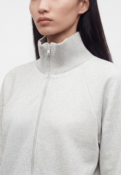 Femme aux longs cheveux foncés portant un pull zippé gris clair avec un col haut côtelé sur un fond uni.